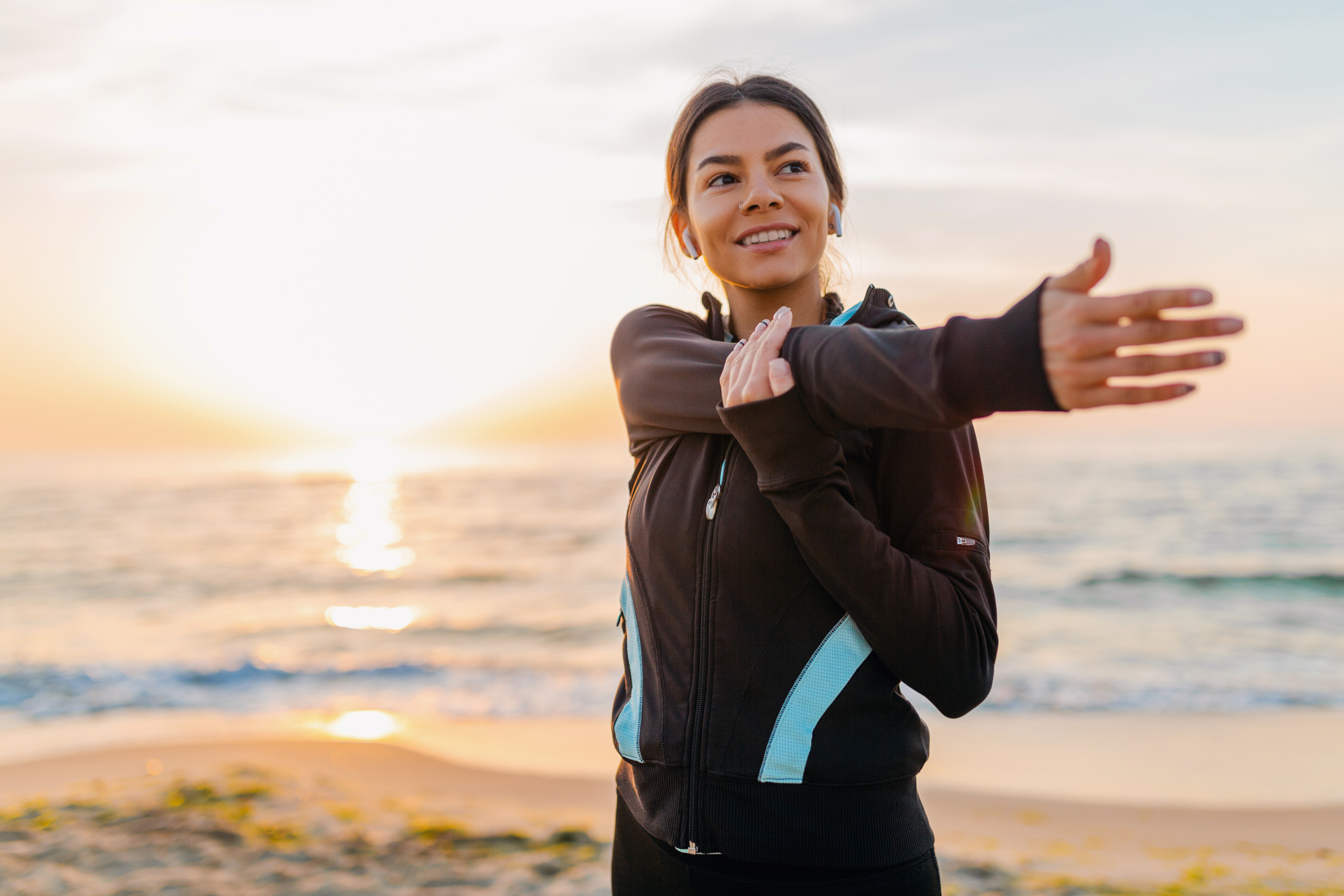 young attractive slim woman doing sport exercises on morning sunrise beach in sports wear, healthy lifestyle, listening to music on earphones, making stretching for arms | Foto: Reprodução/Freepik