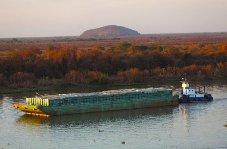 Navio cargueiro na Hidrovia Paraguai-Paraná em trecho de Mato Grosso do Sul, com porto e silos ao fundo