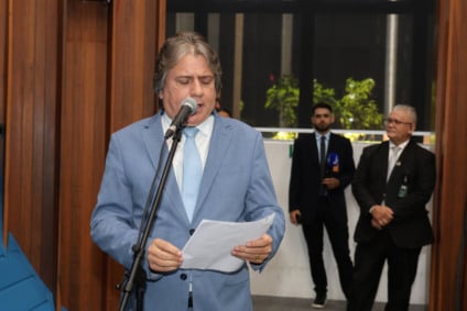 Deputado Caravina falando ao microfone no plenário, com imagem de rodovia ao fundo em telão