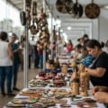 Mãos de artesã com peças artesanais e estandes de feira criativa ao fundo em Mato Grosso do Sul