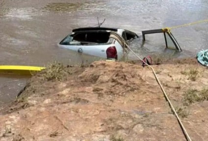 Trecho de rio com carro parcialmente submerso e equipe de resgate ao fundo em cenário rural, sem identificação de pessoas.